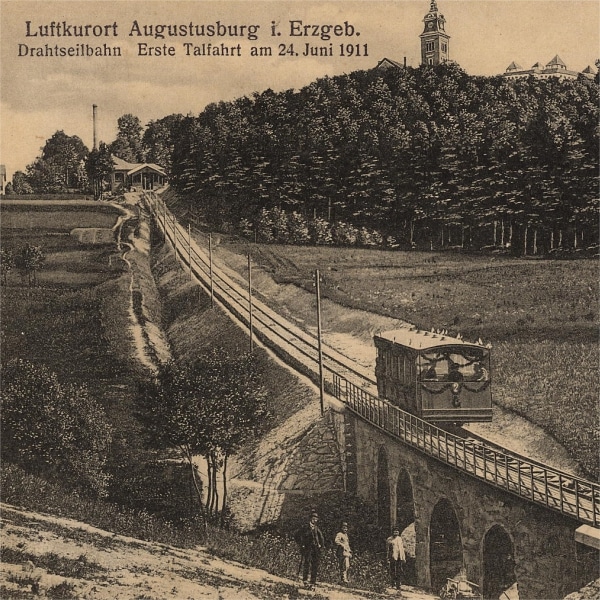 Historische Darstellung, welche die Drahtseilbahn und den oberen Haltepunkt zeigt. Auf dem Motiv ist die Beschriftung "Luftkurort Augustusburg i. Erzgeb. - Drahtseilbahn erste Talfahrt am 24. Juni 1911" vermerkt.