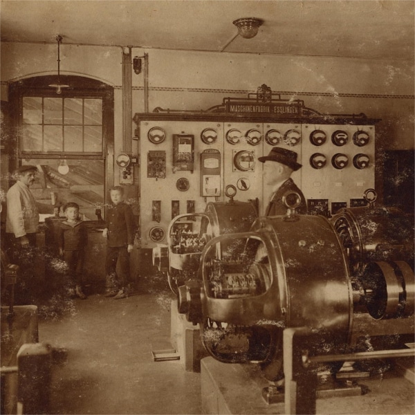 Historisches Foto, das einen der Innenräume eines Haltepunkts der Drahtseilbahn zeigt. Es sind mehrere große Maschinen und vier Personen, davon zwei Kunder zu sehen, die die Gerätschaften betrachten.